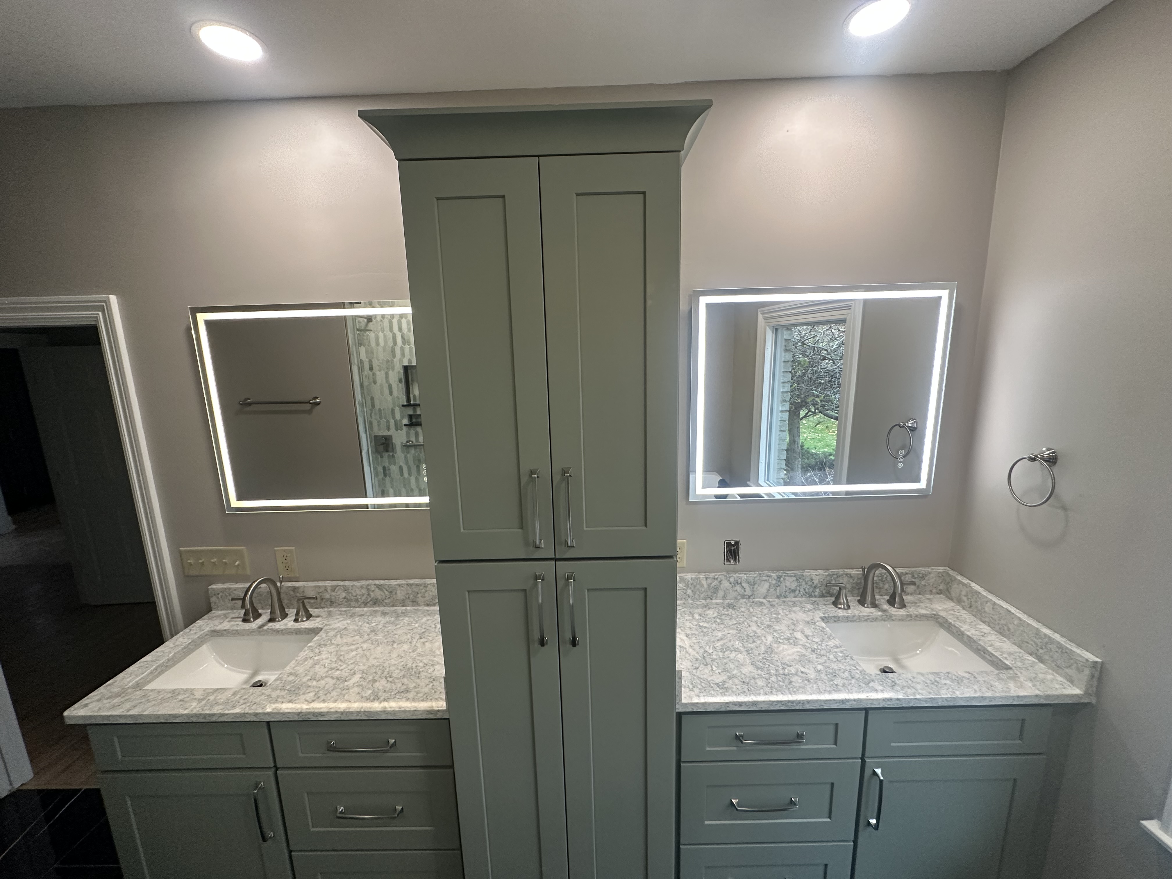 Double vanity with sage green shaker cabinets, a center linen tower, stone-look counters and two backlit mirrors